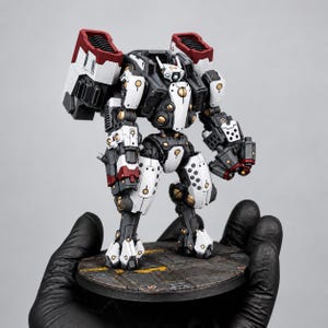 Op de afbeelding: Een miniatuur robotfiguur met een kleurenschema van wit, zwart en donkerrood. Het mechanische figuur heeft een centrale romp, beweegbare ledematen en grote op de schouders gemonteerde stuwraketten. Het staat op een ronde, metalen basis.
