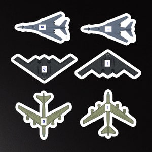 以下が含まれることがあります： さまざまな航空機のデザインが施された6つのステッカー。ステッカーには、グレーとグリーンの濃淡で描かれた軍用機のシルエットが描かれており、それぞれ白い縁取りと青い四角で囲まれた数字が付いています。