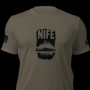 Könnte beinhalten: Hellolivgrünes T-Shirt mit einer schwarzen Grafik eines Messers und dem Wort "NIFE" in fetter, verwitterter Schrift. Auf dem linken Ärmel ist eine amerikanische Flagge aufgedruckt.