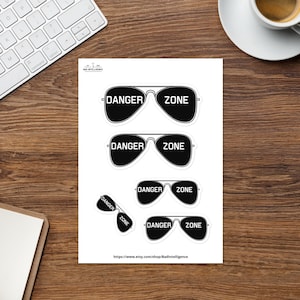 May include: A sheet of white stickers featuring black aviator sunglasses with the words "DANGER ZONE" printed on them. The stickers are in various sizes and orientations. The Etsy shop URL is visible at the bottom.