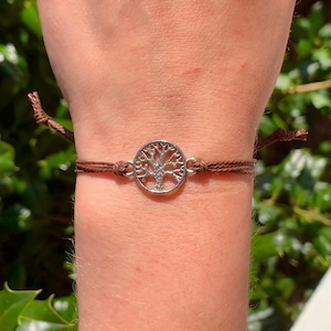 Puede incluir: Pulsera con dije de árbol de la vida plateado y cordón marrón. El dije circular presenta un diseño detallado de árbol. Cierre con nudo corredizo.