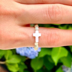 May include: A white cross charm ring with small, pale green beads. The cross is the central feature, set against a blurred backdrop of green leaves and blue flowers. The ring is a delicate piece of jewellery.