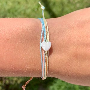 May include: A multi-strand bracelet featuring pastel-colored cords in blue, pink, yellow, and tan. A white heart-shaped bead is centered on the bracelet. The bracelet is tied with a knot closure.