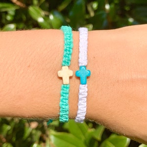 May include: Two friendship bracelets with cross charms. One bracelet is white and the other is turquoise.