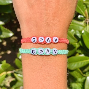 May include: Two friendship bracelets with the words "G>AV" on them. The bracelets are made of colorful string and have white beads with black lettering. The bracelets are on a person's wrist.