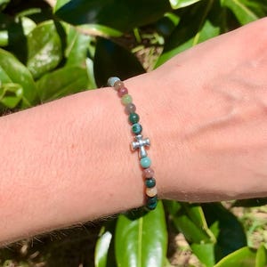 May include: A beaded bracelet featuring a silver cross charm. The bracelet is made of small, colorful beads in shades of green, red, and blue. The cross is centered on the bracelet.