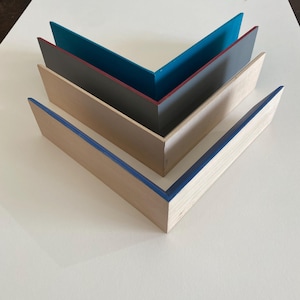 Può includere: Quattro cornici in legno a forma di V, ciascuna con un accento di colore diverso. La cornice superiore ha un interno turchese, la seconda un interno grigio scuro con un bordo rosso e la cornice inferiore ha un bordo blu. Le cornici sono disposte su una superficie bianca.
