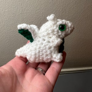 May include: A small white crocheted dragon with green wings. The dragon has green eyes and is sitting in a hand.