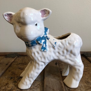 May include: White ceramic planter shaped like a lamb with blue and white ribbon around its neck. The planter has a distressed look with some of the paint chipped off.