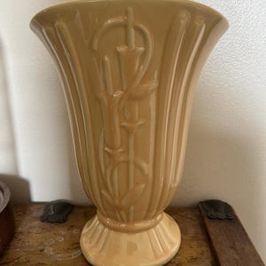 May include: A pale yellow ceramic vase with a fluted design and a floral motif. The vase has a wide mouth and a narrow base.