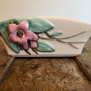 May include: A white ceramic planter with a raised floral design. The planter features a pink flower with green leaves and brown branches.