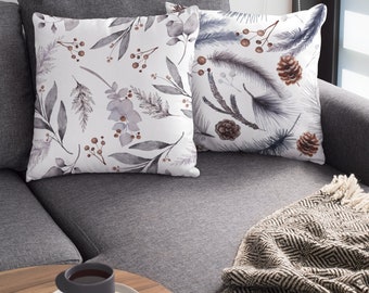 May include: Two decorative cushions with botanical designs. One cushion features a pattern of grey leaves, berries, and flowers. The other cushion has a design of pine needles, pine cones, and berries. The cushions are on a grey sofa.