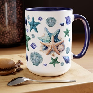 Pode incluir: Caneca de cerâmica branca com interior e alça azuis. A caneca é decorada com ilustrações em aquarela de estrelas do mar e conchas em tons de azul, verde e bege. A caneca está sobre uma superfície de madeira.