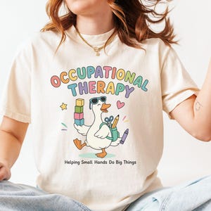 May include: A cream-colored short-sleeve t-shirt featuring a graphic of a white duck wearing sunglasses and a backpack. The text reads OCCUPATIONAL THERAPY and Helping Small Hands Do Big Things, accompanied by illustrations of colorful blocks, a pencil, and scissors.