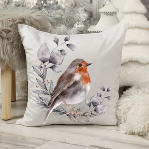 May include: A square, white decorative pillow featuring a watercolor illustration of a robin bird perched on a branch with gray leaves. The bird has a red breast and brown and gray feathers. The pillow is a home decor item.