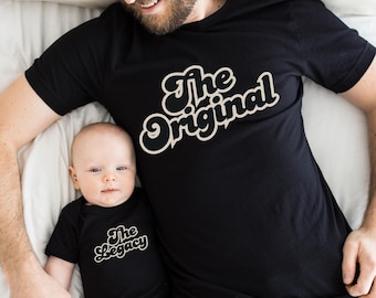 Może przedstawiać: Czarny t-shirt z napisem "The Original" w stylu retro. Obok koszulki znajduje się dziecko w czarnym body z napisem "The Legacy" w pasującym kroju pisma. Widok z góry.