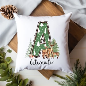 Op de afbeelding: Wit vierkant kussen met een kerstthema. De letter "A" is gevuld met een groen patroon van bomen en sneeuwvlokken. Het kussen toont een familie van herten en de naam "Alexander" in zwart schrift.