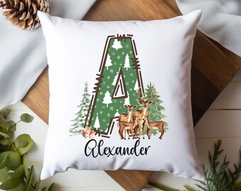 May include: White square pillow with a Christmas theme. The letter "A" is filled with a green pattern of trees and snowflakes. The word "Alexander" is written in black script below a watercolor illustration of deer and trees.