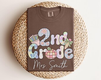 May include: A brown t-shirt with the words "2nd grade" in a colorful, patterned design, along with a graphic of a pencil and apple. The name "Mrs. Smith" is written below. The shirt is folded on a woven mat.