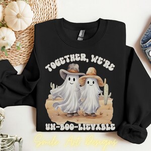 Peut inclure: Sweat-shirt noir avec un motif de deux fantômes portant des chapeaux de cow-boy, dans un décor désertique avec des cactus. Le texte "TOGETHER, WE'RE UN-BOO-LIEVABLE" est imprimé au-dessus et en dessous des fantômes. Design par Smile Art Designs.