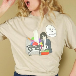 K&ouml;nnte beinhalten: Beiges T-Shirt mit einer Grafik einer Person und einer Kuh, mit dem Text "You are my begonias!". Die Person tr&auml;gt eine Sonnenbrille und eine blaue Jeans.
