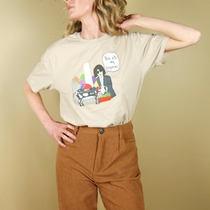 K&ouml;nnte beinhalten: Beiges T-Shirt mit einer grafischen Illustration, die eine Person im Anzug, eine Kuh und den Text "You are my begonias!" zeigt. Das Shirt wird mit einer braunen Cordhose kombiniert.