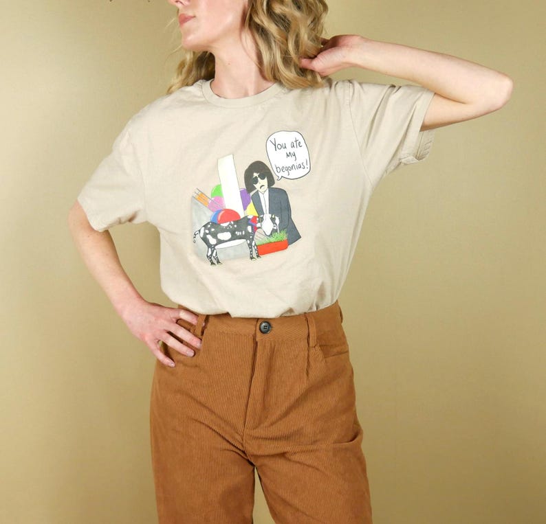 K&ouml;nnte beinhalten: Beiges T-Shirt mit einer grafischen Illustration, die eine Person im Anzug, eine Kuh und den Text "You are my begonias!" zeigt. Das Shirt wird mit einer braunen Cordhose kombiniert.