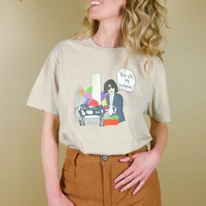 K&ouml;nnte beinhalten: Beiges T-Shirt mit einer Cartoon-Illustration mit einer Kuh, einer Person im Anzug und dem Text "You are my begonias!". Das Shirt hat einen Rundhalsausschnitt und kurze &Auml;rmel.