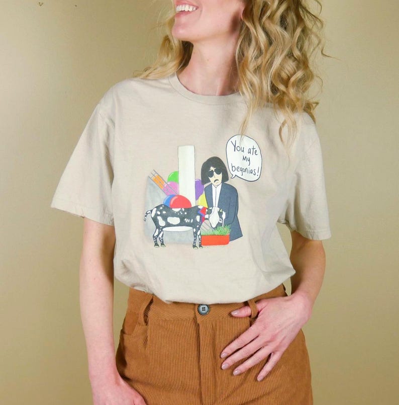 K&ouml;nnte beinhalten: Beiges T-Shirt mit einer Cartoon-Illustration mit einer Kuh, einer Person im Anzug und dem Text "You are my begonias!". Das Shirt hat einen Rundhalsausschnitt und kurze &Auml;rmel.