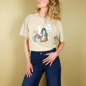 K&ouml;nnte beinhalten: Beiges T-Shirt mit einer Cartoon-Illustration einer Person mit Sonnenbrille, einer Kuh und dem Text "You are my begonias!". Die Person tr&auml;gt einen Anzug und h&auml;lt einen Blumenkorb. Das Shirt wird mit einer blauen Jeans kombiniert.