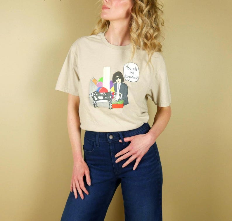 K&ouml;nnte beinhalten: Beiges T-Shirt mit einer Cartoon-Illustration einer Person mit Sonnenbrille, einer Kuh und dem Text "You are my begonias!". Die Person tr&auml;gt einen Anzug und h&auml;lt einen Blumenkorb. Das Shirt wird mit einer blauen Jeans kombiniert.