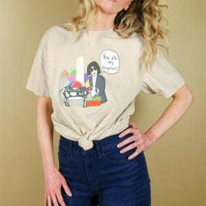 K&ouml;nnte beinhalten: Beiges T-Shirt mit einer Cartoon-Illustration einer Ziege, einer Person im Anzug und dem Text "You ate my begonias!". Die Grafik enth&auml;lt bunte Formen und einen Blumentopf. Das T-Shirt ist an der Taille geknotet.