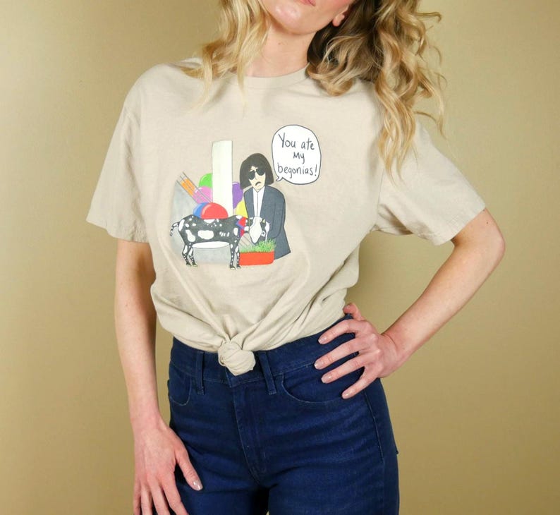 K&ouml;nnte beinhalten: Beiges T-Shirt mit einer Cartoon-Illustration einer Ziege, einer Person im Anzug und dem Text "You ate my begonias!". Die Grafik enth&auml;lt bunte Formen und einen Blumentopf. Das T-Shirt ist an der Taille geknotet.