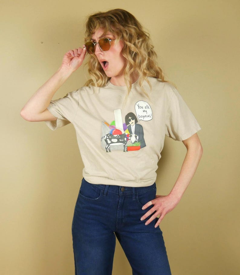 K&ouml;nnte beinhalten: Beiges T-Shirt mit einer Grafik einer Person und einer Kuh, mit dem Text "You are my begonias!". Die Person tr&auml;gt eine Sonnenbrille und eine blaue Jeans.