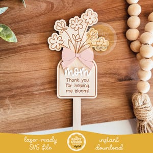 May include: A laser-cut wooden sign with a pink bow and a bouquet of flowers. The sign reads "mom Thank you for helping me bloom!"