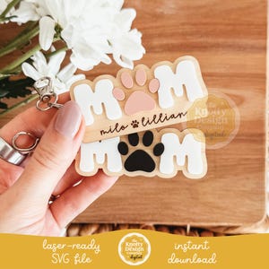 May include: Wooden keychain with the word "MOM" in white letters, featuring a paw print design in pink and black. The keychain includes the names "milo" and "lillian". The design is laser-ready and available for instant download.
