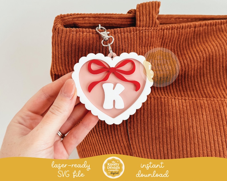 May include: A white heart-shaped keychain with a red bow and the letter K in the center. The keychain is attached to a brown corduroy bag.