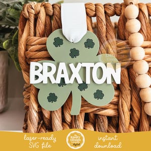 May include: A green shamrock-shaped ornament with the name "BRAXTON" in white letters. The ornament is hanging from a wicker basket.