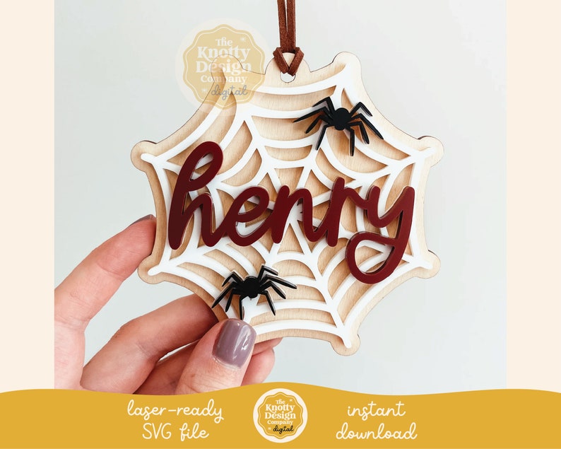 Pu&ograve; includere: Un ornamento in legno a forma di ragnatela con il nome "henry" in lettere rosse. La ragnatela &egrave; bianca e ha tre ragni neri. L'ornamento &egrave; appeso a uno spago marrone.