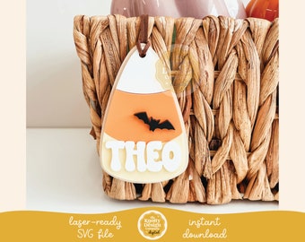 Può includere: Un'etichetta a forma di caramella di mais a tema Halloween con il nome "THEO" in lettere bianche. Un pipistrello nero è centrato sulla sezione arancione. L'etichetta è attaccata a un nastro marrone.
