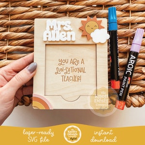 May include: A wooden frame with a cutout for a photo, featuring a sun and cloud design. The frame has a personalized message that reads "Mrs. Allen You Are A Sun-Sational Teacher!" and a rainbow design at the bottom. The frame is accompanied by two oil-based paint pens.