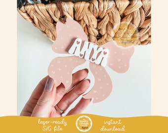 Könnte beinhalten: Eine rosa schleifenförmige, laserfertige SVG-Datei mit dem Namen "ANYA" in weißen Buchstaben. Die Schleife ist mit kleinen Herzformen verziert. Ein brauner Faden ist an der Schleife befestigt. Das Bild enthält auch den Text "sofortiger Download".
