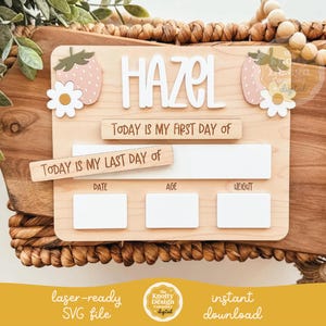Puede incluir: Tablero de hitos de madera con el nombre "HAZEL" en letras blancas. El tablero presenta detalles de fresas y margaritas, y espacio para registrar "DATE", "AGE" y "WEIGHT". El texto dice "TODAY IS MY FIRST DAY OF" y "TODAY IS MY LAST DAY OF".