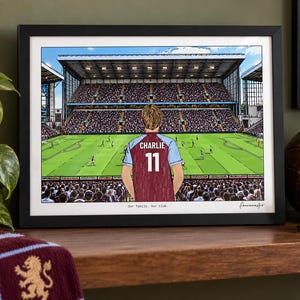 May include: A framed horizontal print depicting a soccer stadium view from behind a player wearing a claret and blue jersey with the name CHARLIE and number 11 on the back. The text Our family. Our club. is printed at the bottom of the illustration.