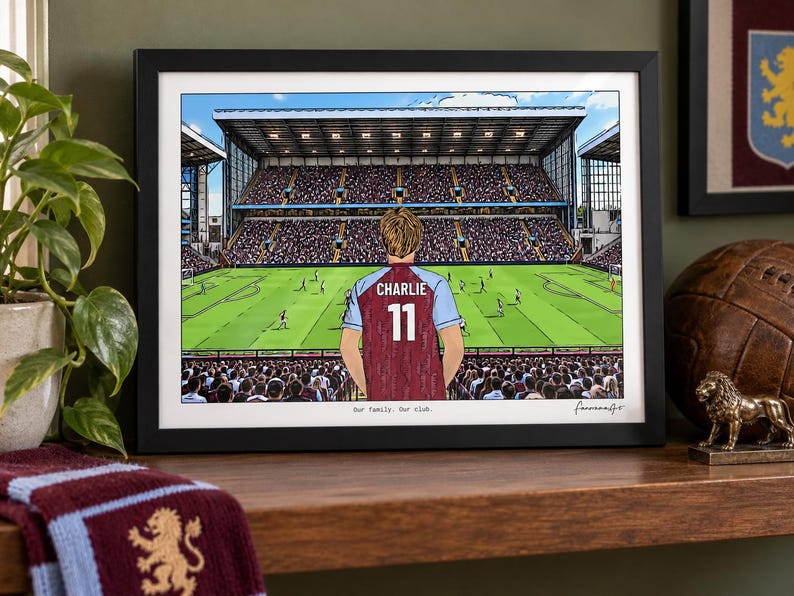 May include: A framed horizontal print depicting a soccer stadium view from behind a player wearing a claret and blue jersey with the name CHARLIE and number 11 on the back. The text Our family. Our club. is printed at the bottom of the illustration.