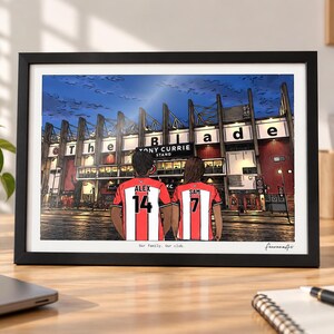 May include: A framed digital illustration of two people wearing red and white striped soccer jerseys with the names ALEX 14 and SAM 7 on the back, standing before a stadium with a sign reading The Blade and Tony Currie Stand.
