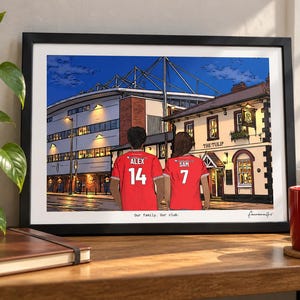 Pu&ograve; includere: Un'illustrazione incorniciata di due persone che indossano maglie da calcio rosse con i nomi ALEX 14 e SAM 7 sul retro, davanti a uno stadio e a un pub chiamato THE TULIP. La stampa include il testo Our family. Our club. in basso.