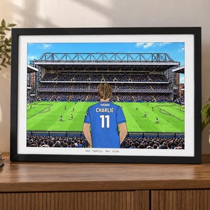 May include: A framed illustration of a soccer stadium with a person wearing a blue jersey with the name CHARLIE and the number 11 on the back. The text Our family. Our club. is printed at the bottom of the print inside a black frame.