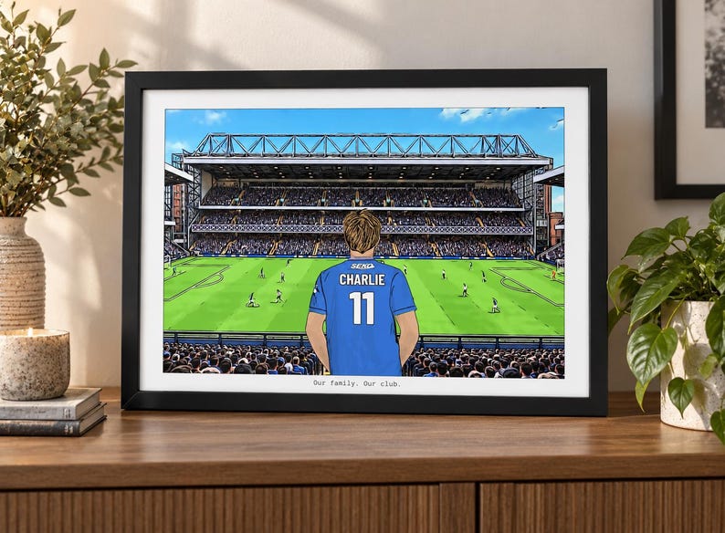 May include: A framed illustration of a soccer stadium with a person wearing a blue jersey with the name CHARLIE and the number 11 on the back. The text Our family. Our club. is printed at the bottom of the print inside a black frame.