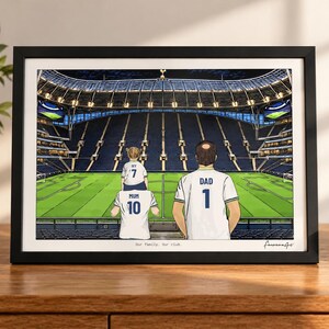 Puede incluir: Una ilustraci&oacute;n enmarcada de una familia de tres personas dentro de un gran estadio de f&uacute;tbol. Las figuras visten camisetas blancas con el texto MUM 10, IVY 7 y DAD 1. La impresi&oacute;n incluye el texto Our family. Our club. en un marco negro.